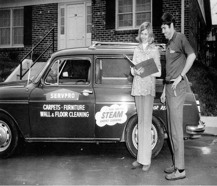 A black and white photo with a SERVPRO vehicle in the center and a SERVPRO professional with a woman on the right.