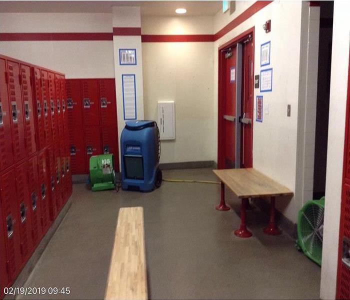 Locker room with red lockers to the left and red doors to the right. Dehumidifiers are located on the floor.